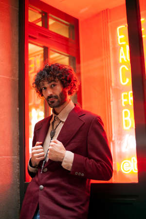 Fashionable young Caucasian man in a red jacket looking at the camera while adjusting lapels with both hands. An orange neon light illuminates his face. Millennial with a beard.の写真素材