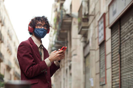 Happy millennial man smiling with mask after lock reopening - Hipster man with glasses enjoying with smart phone in hands and headphones on a Barcelona street. copy spaceの写真素材