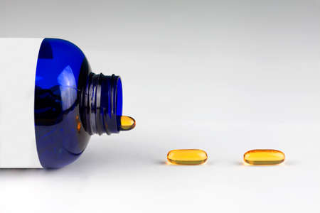 Close up of fish oil supplement capsules coming out of semitransparent blue plastic bottle on white background. Selective focus and copy space. Healthy diet conceptの写真素材