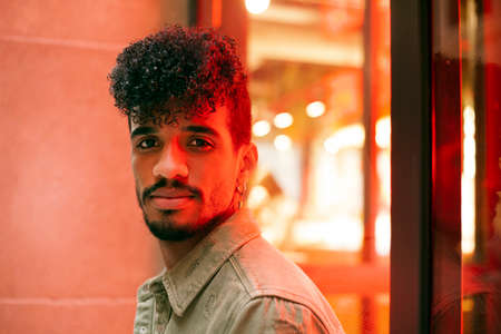 Portrait of young biracial curly haired man with orange neon light reflection on face. Looking towards the camera. Millennial with beard and earring. Horizontal image.の写真素材