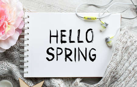 A notebook with the inscription HELLO SPRING on a light table along with a pink flower and earphones for the phone. Natural light. Marketing concept. Top view, flat lay. Spring theme.の写真素材
