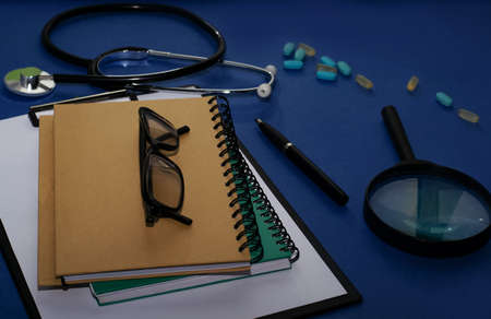 Doctor's workplace. Top view on a blue background with medical equipment - stethoscope, tablet with a blank sheet of paper, glasses. medical concept.の写真素材