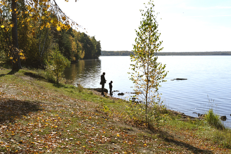View to a lake in autum and silhouettes of man and boy walking around. Father and son on a lake shore on a sunny day of autumn.の写真素材