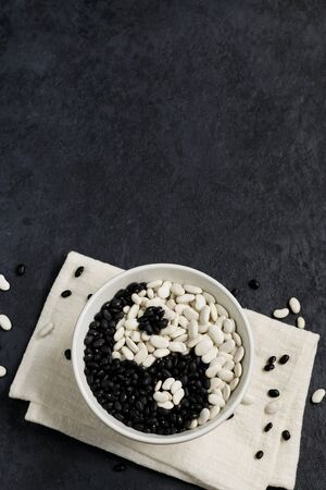 Beans on a dark background. White beans and black beans in a gray bowl on a napkin. Sign Yin Yang. Top view. Vertical position. Copy spaceの写真素材