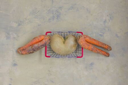 An ugly heart with potatoes and two ugly carrots on a light background in a shopping basket. Funny, unusual concept of vegetables or food waste. top view. Horizontal orientation.の写真素材