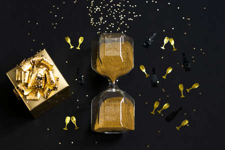 Golden christmas flat lay with confetti, gift box and hourglass on black background. New Year...