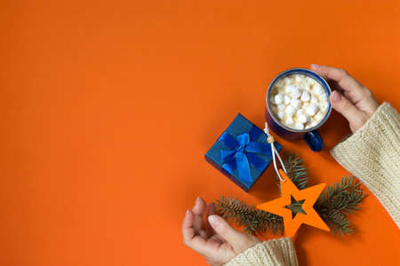 Female hands hold a blue gift box and a hot drink with marshmallows on an orange background....