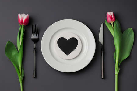 Creative table setting with hearts on a white plate, black fork and knife, tulips on a dark...