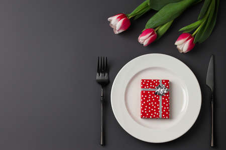 Creative table setting with gift box on a white plate, black fork and knife, tulips on a dark background. Valentine's Day, Wedding Day, Birthday, Women's Day and Mother's Day. Flat lay. Copy space.の写真素材