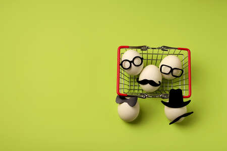 Group of white eggs in black hats and glasses in a shopping basket on a green background. Easter holiday shopping minimal concept. Flat lay. Copy spaceの写真素材