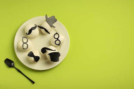 A group of white eggs in black hats and glasses on a plate on a green background. Festive breakfast. Creative minimal concept for Easter. Flat lay. Copy spaceの写真素材