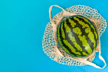 Watermelon in a string bag on a blue background. Healthy food delivery concept. Detox in a reusable eco mesh. Copy spaceの写真素材