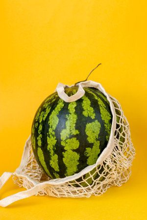 Watermelon in a string bag on a yellow background. Detox in a reusable eco mesh. Healthy food delivery conceptの写真素材