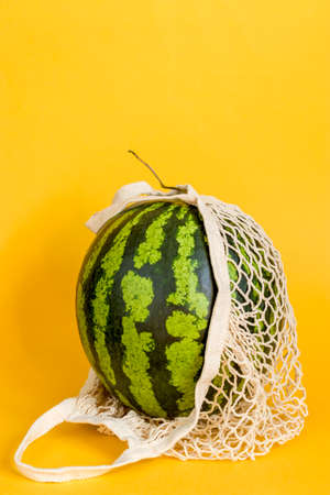 Watermelon in a string bag on a yellow background. Detox in a reusable eco mesh. Healthy food delivery concept. Copy spaceの写真素材