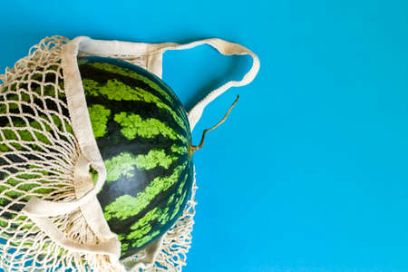 Watermelon in a string bag on a blue background. Detox in a reusable eco mesh. Healthy food delivery concept. Copy spaceの写真素材