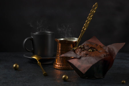 Cup of brewed coffee in turk with fresh sweet chocolate muffin on black background. Low keyの写真素材