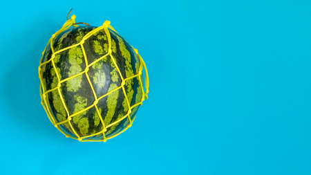 Watermelon in a string bag on a blue background. Healthy food delivery concept. Detox in a reusable eco mesh. Copy spaceの写真素材