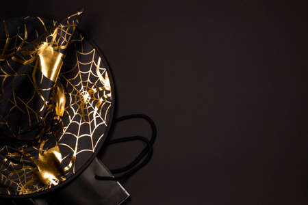 Halloween shopping holiday. Black witch hat with spider web and spider pattern, shopping bag on black background, flat lay. Copy spaceの写真素材