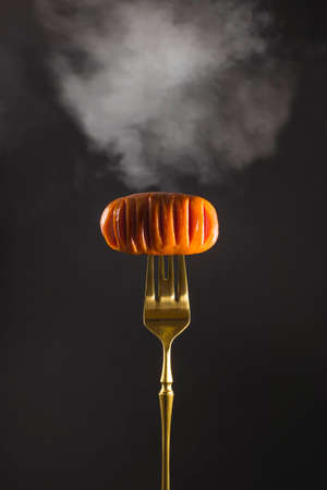 Delicious hot grilled meat sausage with smoke on a golden fork on a black background. Vertical image with copy space.の写真素材