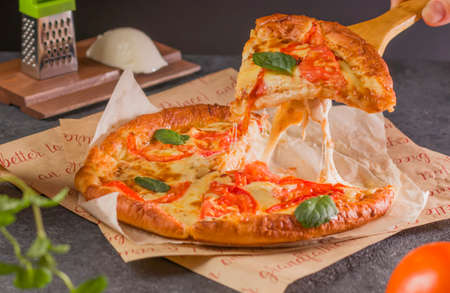 Fresh delicious fast food. A piece of hot pizza with melting cheese on the table. Traditional Margherita pizza with tomatoes, basil and mozzarella cheese close-up on a dark background.の写真素材