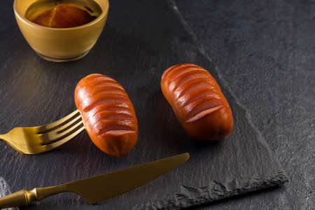 Delicious hot grilled meat sausages on a slate board. Gold colored table knife and fork on a black...