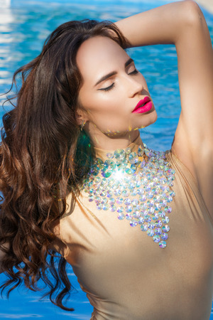 Beautiful brunette in golden swimsuit standing in swimming pool enjoying the sun and blue waterの写真素材