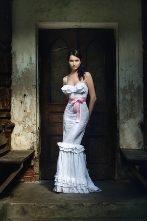 Elegant woman in a stylish long white gown posing indoors in front of a dark closed door with her hands clasped behind her back and a serious expressionの写真素材