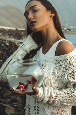 Beautiful girl  with dark hair standing with golden fish near the beach, concept of loving nature and animalsの写真素材