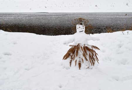 A sad snowman in a cane skirt stands on the river bank on a winter day.の写真素材