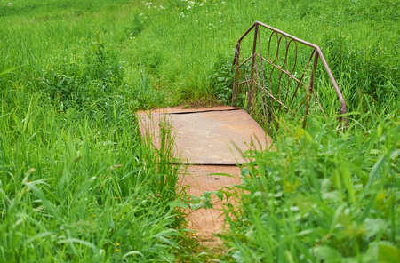 A rusty metal pedestrian bridge spanning a grassy stream.の写真素材