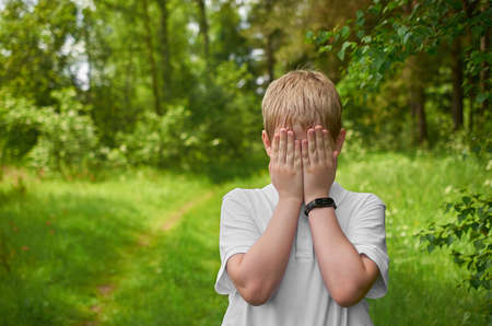 A European preteen in a white T-shirt covered his face with his hands in a summer forestの写真素材