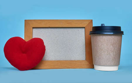 A paper cup for coffee, a toy heart and a wooden frame with a place for text on a blue background.の写真素材