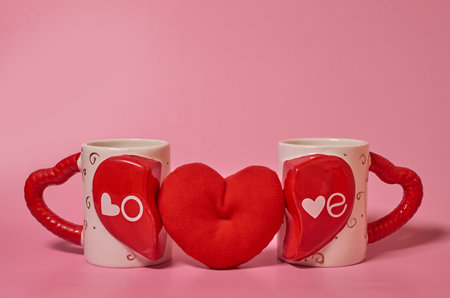 Two paired mugs with the inscription Love, handles in the form of hearts and a red plush heart between them on a solid pink backgroundの写真素材
