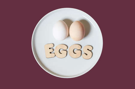 The word Eggs is made of wooden letters and two chicken eggs on a round white plate on a colored...