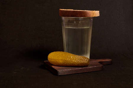 A faceted glass of vodka, rye bread and a pickled cucumber on a wooden stand. Still life on a dark backgroundの写真素材