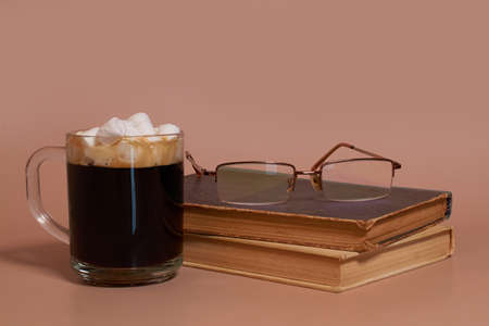 A glass cup of coffee with marshmallows stands next to a stack of old books and glasses on a colored background. Side view, space for text, nobodyの写真素材