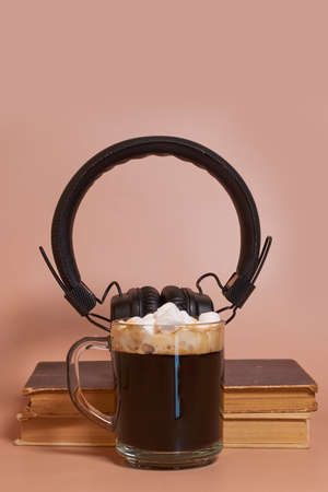 A glass cup of coffee with marshmallows stands next to a stack of old books and headphones on a...
