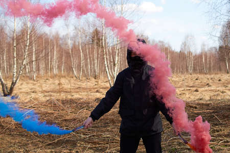 A preteen in a black jacket and a mask covering his face holds two colored smoke bombs. portrait. horizontal formatの写真素材