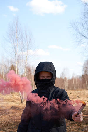 A preteen in a black jacket and a mask covering his face holds a checker with colored smoke. vertical formatの写真素材