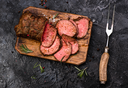 Sliced grilled roast beef with fork for meat on wooden cutting board. Black background. Top view.の写真素材