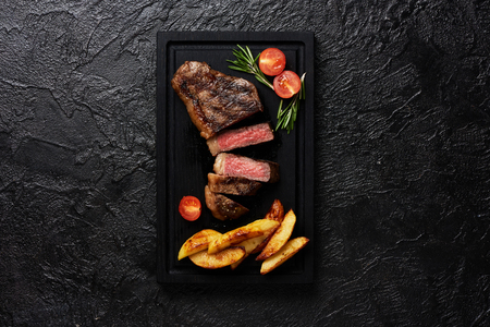 Meat Picanha steak, traditional Brazilian cut with tomatoes, potato wedges and rosemary on black meat cutting board with copy space. Black concrete background.の写真素材