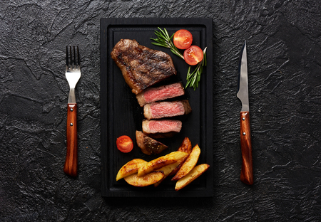 Meat Picanha steak, traditional Brazilian cut with tomatoes, potato wedges and rosemary on black meat cutting board. Steak with fork and knife. Black concrete background.の写真素材