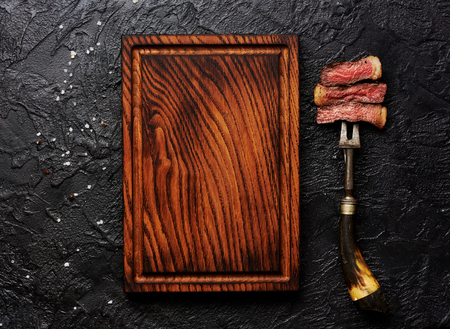 Slices of medium rare grilled Steak on meat fork on black concrete background. Copy space. Wooden meat cutting board. Top view.の写真素材