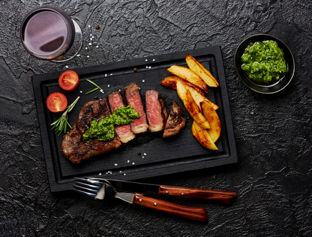 Meat Picanha steak, traditional Brazilian cut with potato wedges, chimichurri sauce and rosemary on black meat cutting board. Steak and a glass of red wine with fork and knife.の写真素材
