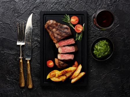 Meat Picanha steak, traditional Brazilian cut with potato wedges, chimichurri sauce and rosemary on black meat cutting board. Steak and a glass of red wine with fork and knife.の写真素材