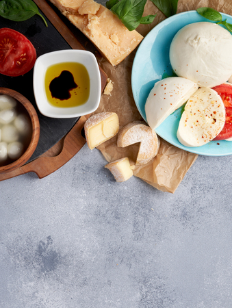 Italian cheeses. Mozzarella Buffalo, crottin, balls, Stracciatella, basil leaves, tomatoes, olive oil with vinegar on gray background. Top view, copy space.の写真素材
