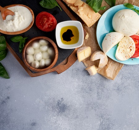 Italian cheeses. Mozzarella Buffalo, crottin, balls, Stracciatella, basil leaves, tomatoes, olive oil with vinegar on gray background. Top view, copy space.の写真素材