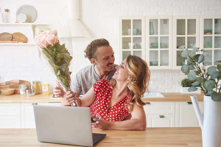 Happy family couple in modern kitchen with laptop and coffee. A man in love gives flowers to a woman.の写真素材
