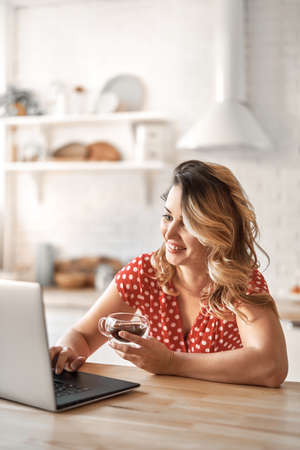 Happy beautiful woman drinking coffee. Woman working on a laptop in kitchen.の写真素材