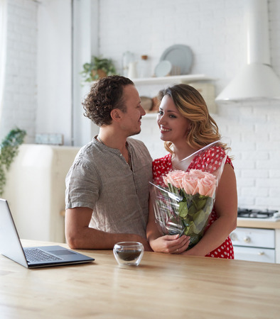 Summer holidays, love, relationship - couple with bouquet of flowers in the kitchen. Laptop on the table.の写真素材